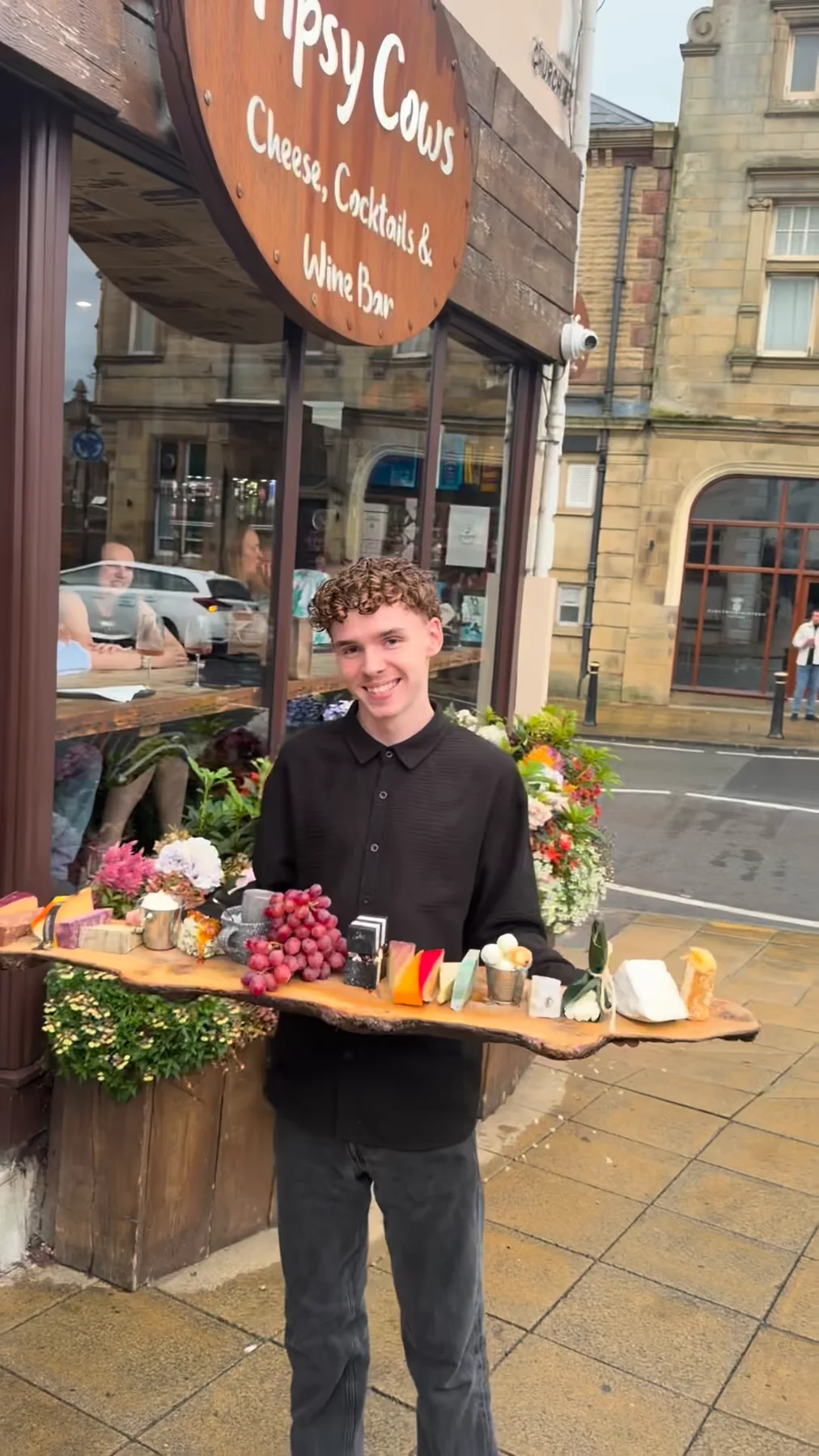 A member of the Tipsy Cows team holding a long cheese board outside the Church Street shopfront