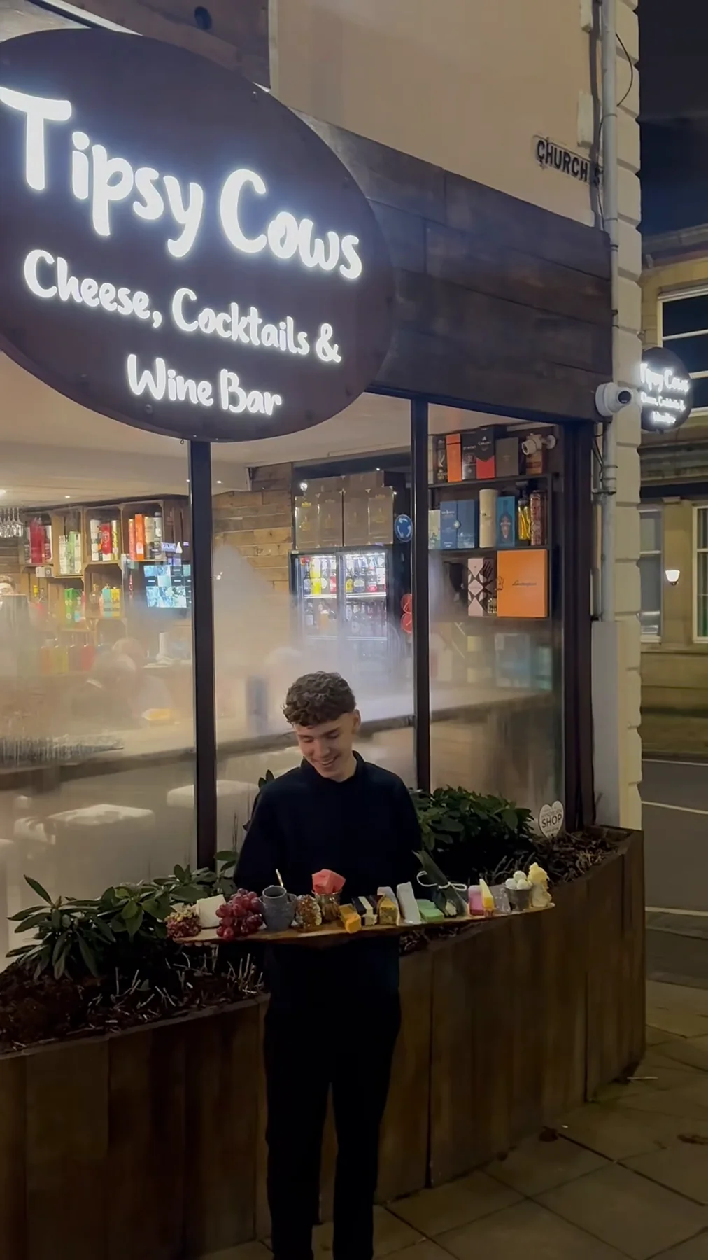 A member of the team holding a cheese board outside the lit Tipsy Cows shopfront